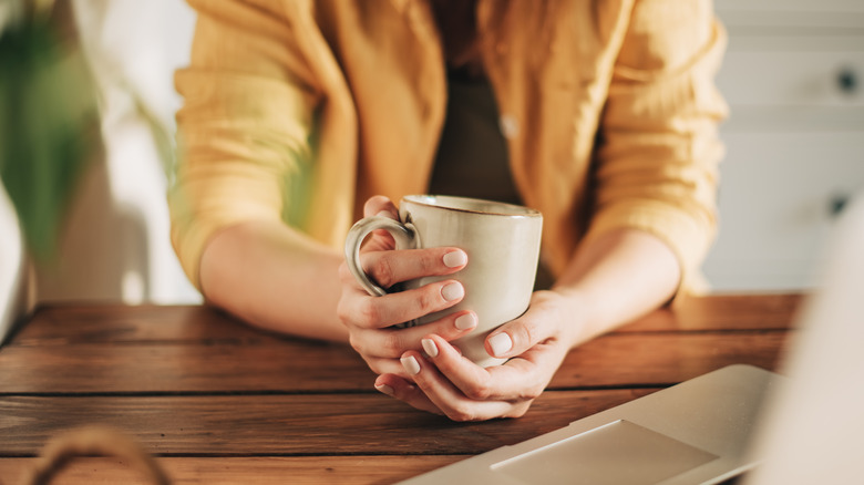 A person holding a stoneware mug in their hands on top of a wooden table.