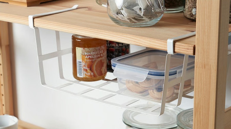 PÅLYCKE white metal clip-on basket hung on kitchen cabinet shelf, holding jars and container.