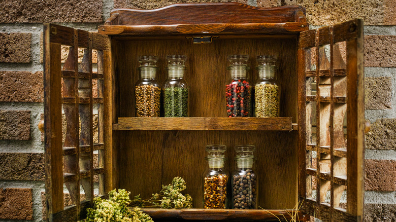 A wooden spice rack with doors hanging on a brick wall with spice-filled jars on the shelves.