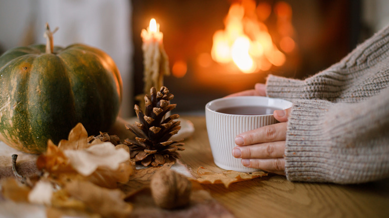 Beginnings of a centerpiece with a pumpkin, pine cones, and leaves in front of a fire with a cup of coffee or hot cocoa