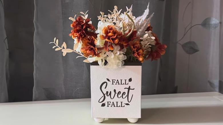 White box on raised feet with "fall sweet fall" painted on it holding fall colored flowers