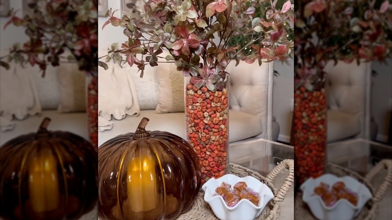 Glass pumpkin next to vase filled with mini pumpkin pods and flowers on a table with bowl of caramels