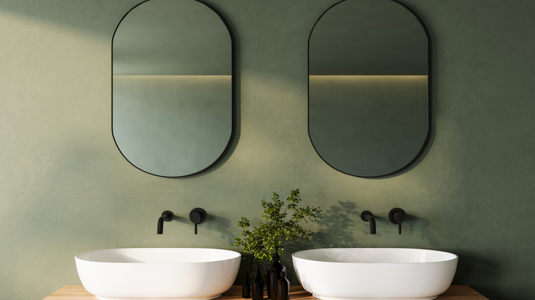A bathroom vanity with two over mounted sinks on a sage green wall.