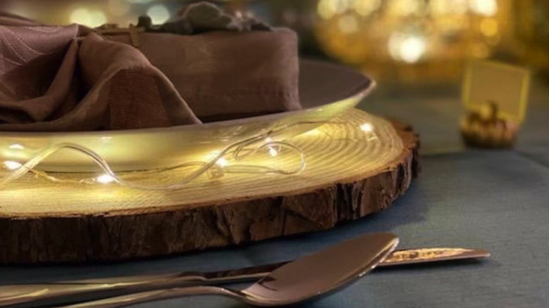 Plate swith a fancy napkin sitting on a thin sliced wood place mat with fairy lights on it and silverware resting on the table near it