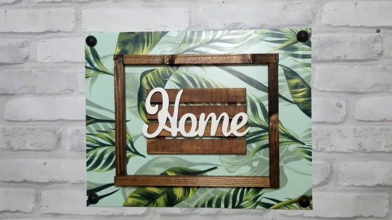Wall decor with a tropical leaf background, a wooden frame, and a mini wood pallet in the middle with the word "Home" on it