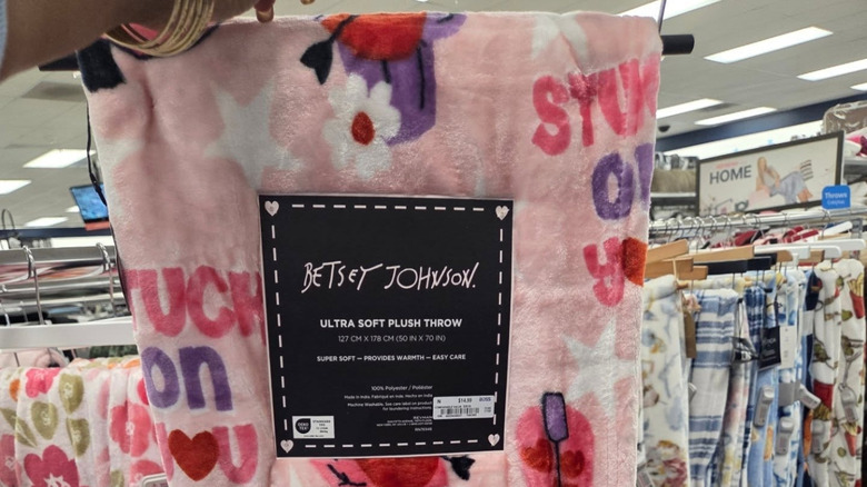 Hand holds up a pink plush throw from Betsey Johnson in front of various other throws