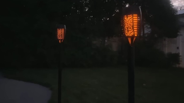 Four faux tiki torches made from solar lights and broom handles line a backyard as string lights hang in a tree.