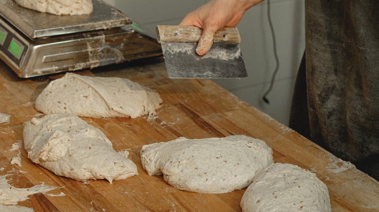 Four dough balls on a wood surface and one on a a scale with a hand holding a bench scraper over the prep area