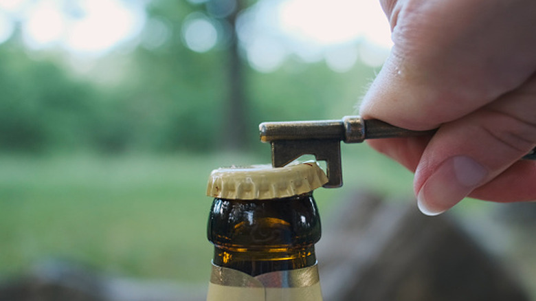 hand using a key-shaped bottle opener to pry a bottle cap off a glass bottle outside