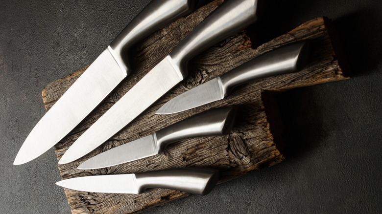 Assortment of stainless steel kitchen kives on a wood cutting board on a dark colored surface