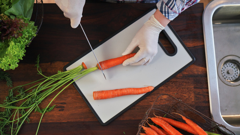 Gloved hands cutting the tops off carrots on a plastic cuttingboard over a wood counter near a sink