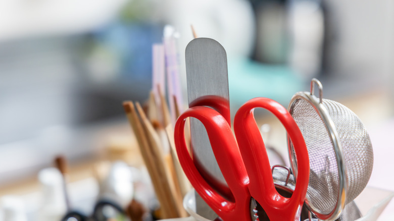 Red Kitchen shears sticking out of a utlility container with a metal frosting knife, a small mesh strainer, and other assorted kitchen utensils