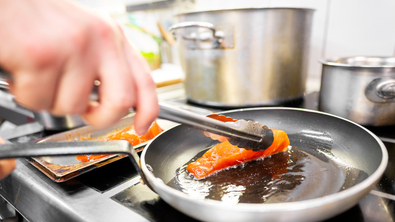 Hand using metal tongs to place raw salmon in hot oil in a metal frying pan
