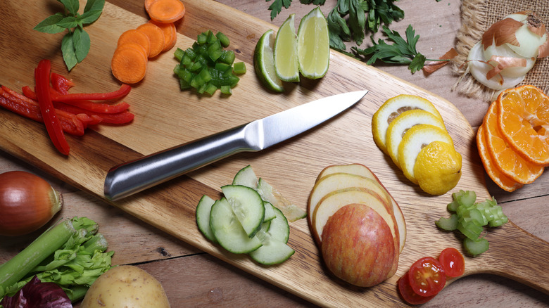 a paring knife sitting on a wood cutting board with various sliced vegetables, fruits, and herbs