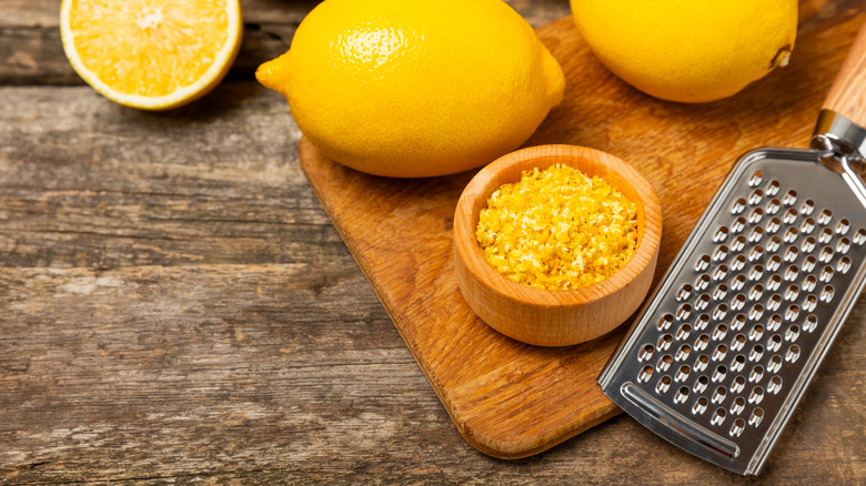 Lemons on a cutting board with a small wood bowl full of lemon zest near a small zester