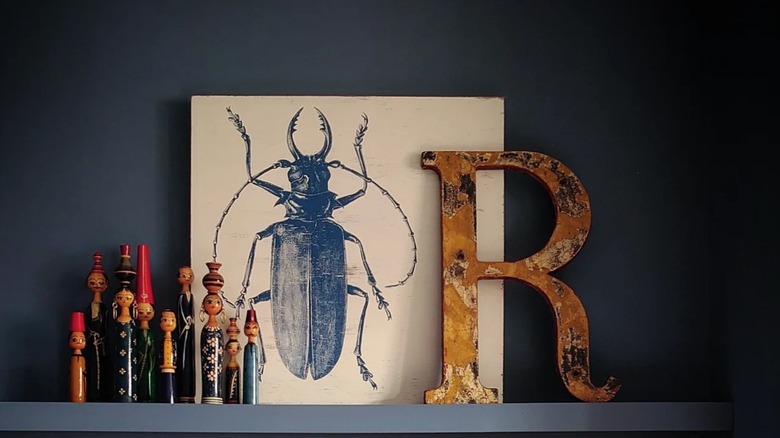 Decorated peg dolls, a cockroach drawing, and a wooden R on a shelf.
