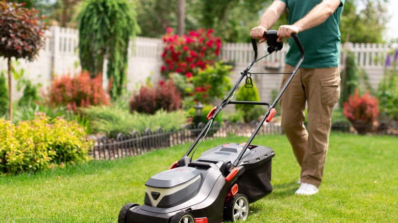 Person in green shirt and brown pants pushing a lawn mower in a yard