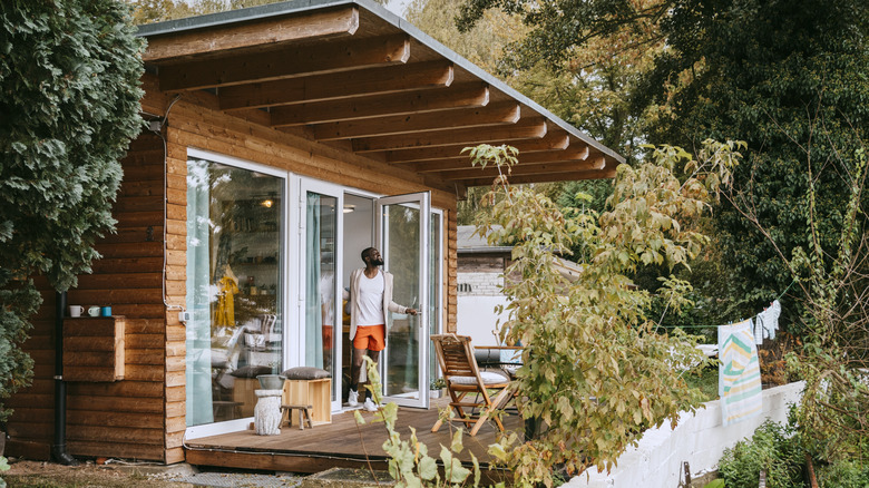 Small wooden home with large windows and a wood porch with bistro set, a man holds open the door