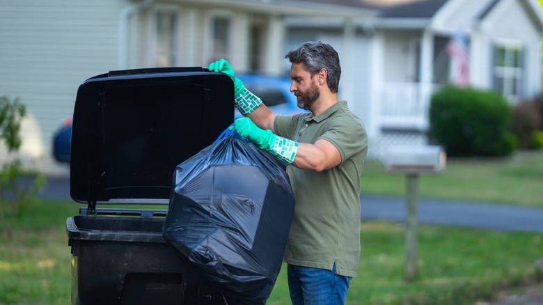 Man wearing rubber gloves throwing trash bag in trash can outside home