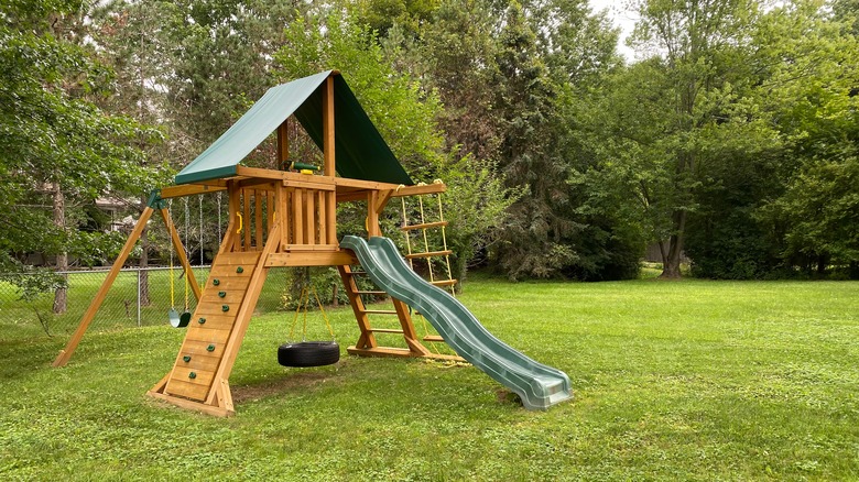 Wooden play structure with a slide, rope ladder, swing set, and climbing wall in a fenced yard