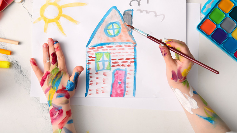 Ppaint-covered kid's hands painting a colorful house on white paper
