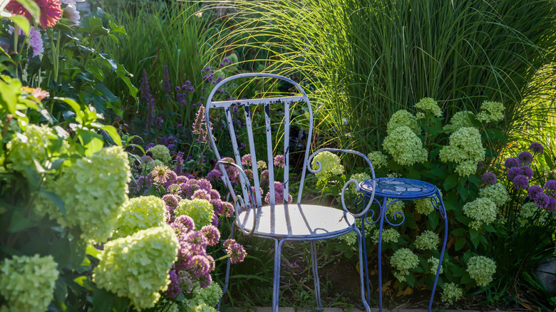 A vintage chair and table in the corner of a garden, surrounded by flowers in dappled sunlight.