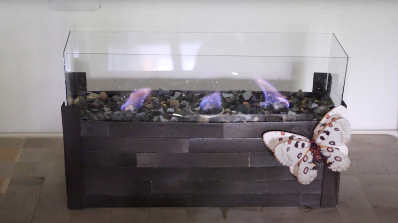 A mini tabletop fire pit made from a wine box, glass panels, and some candles with a decorative butterfly affixed on the side.
