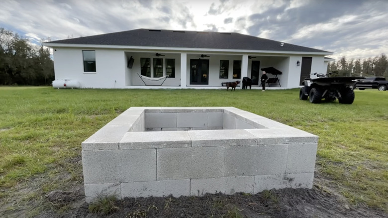 A fire pit made from concrete blocks sits on the lawn in the backyard of a single floor home.