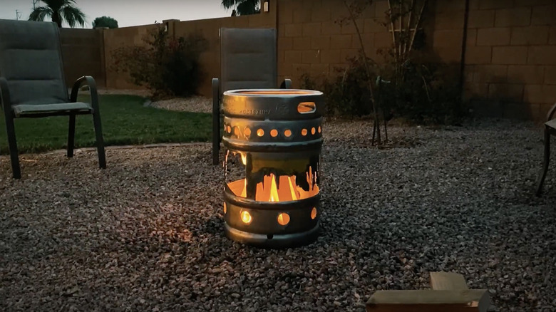 A fire roars inside a beer keg fire pit sitting on a gravel patio in a backyard at sunset.