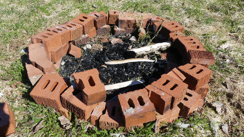 A fire pit made from upcycled and broken red clay bricks sits on a lawn with ash and burned wood inside.