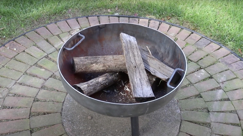 A fire pit made from an old propane tank sits on a cobbled surface and is filled with cut firewood.