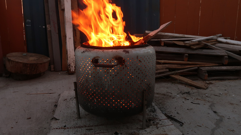 An old washing machine drum turned into a fire pit burns wood on a concrete floor inside a shed.