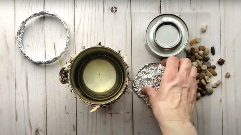A hand grabs a piece of aluminum foil sitting on a wooden table with aquarium stones, tin cans, and other craft supplies.