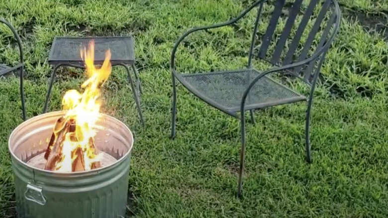 A fire pit made from a trash can sits on a lush lawn next to some metal outdoor furniture.