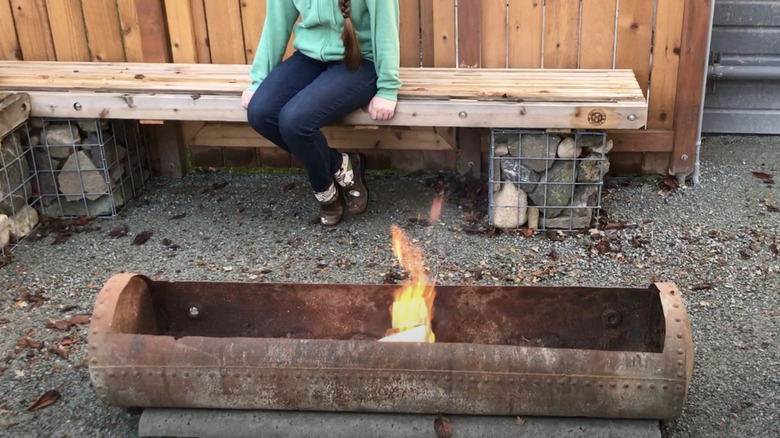 A woman sits on a bench behind a fire pit made from an old hot water heating tank.