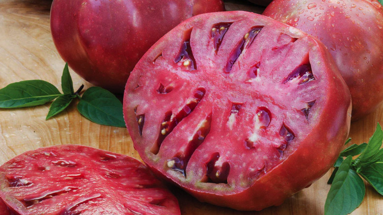 A 'Cherokee Purple' Tomato sliced with basil on a cutting board with two whole 'Cherokee Purple' tomatoes behind it
