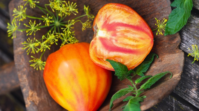 A full 'Orange Oxheart' tomato next to one cut in half on a wood plate with leafy garnishments