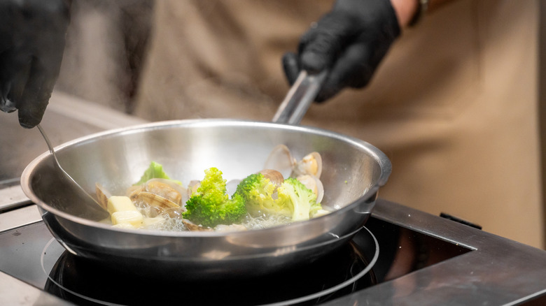 Person wearing black gloves cooking vegetables in stainless steel pan.