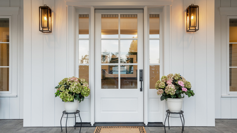 A white front door on a white house is flanked by lanterns and potted hydrangeas