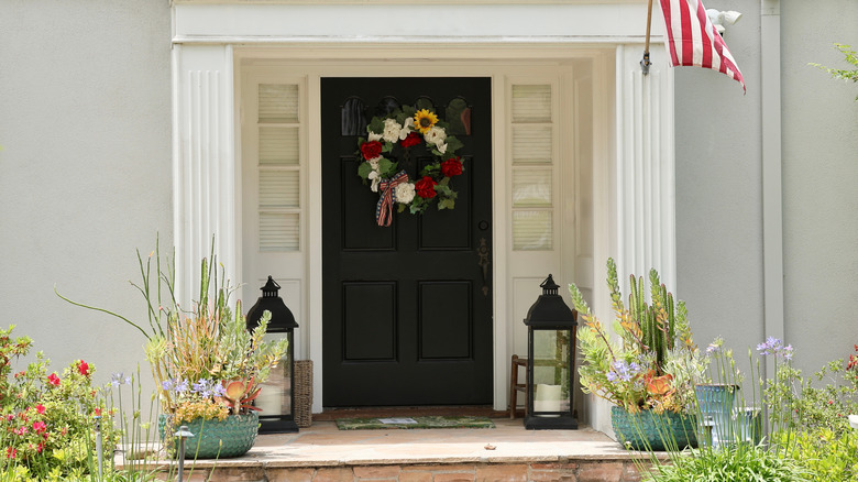 A floral wreath hangs on a black front door surrounded by lanterns and potted plants