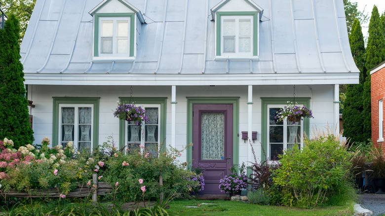 A white cottage with a purple front door is surrounded by lush landscaping