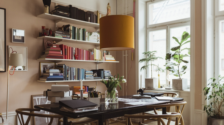 A home office space with lots of natural light, several houseplants, and open shelves filled with color-coordinated books.