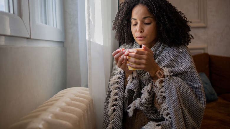 A woman sits beside a radiator holding a mug in her hands.