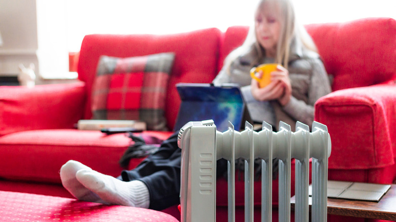 space heater in foreground while elderly woman sits on sofa with mug and laptop