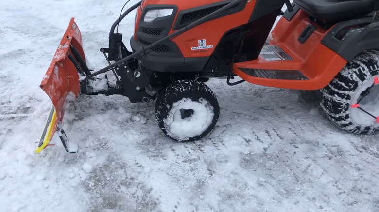 A snow blade attachment in front of a seated lawn mower in cleared snow.
