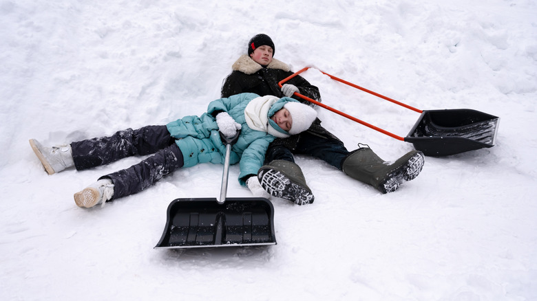 Two exhausted teenagers laying in snow with shovels.