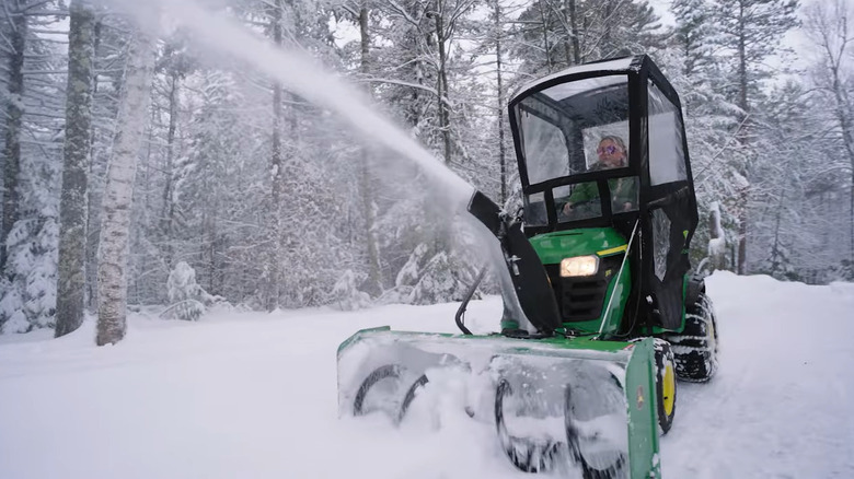 A green John Deere lawn tractor with a snow blower attachment and cab.