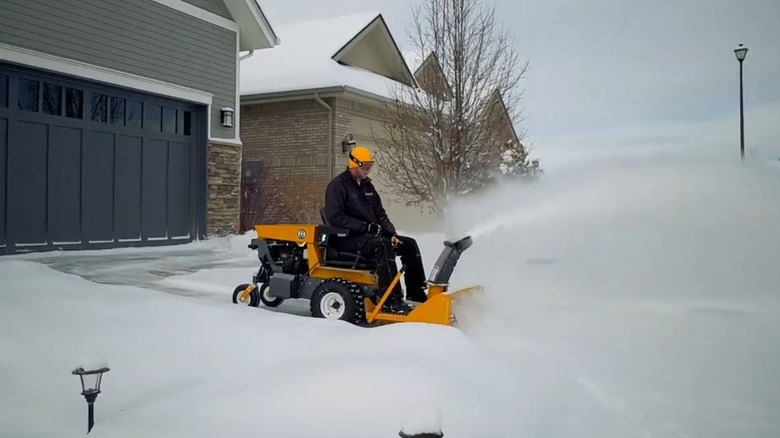 A man riding a yellow lawn mower with a snow thrower attachement.