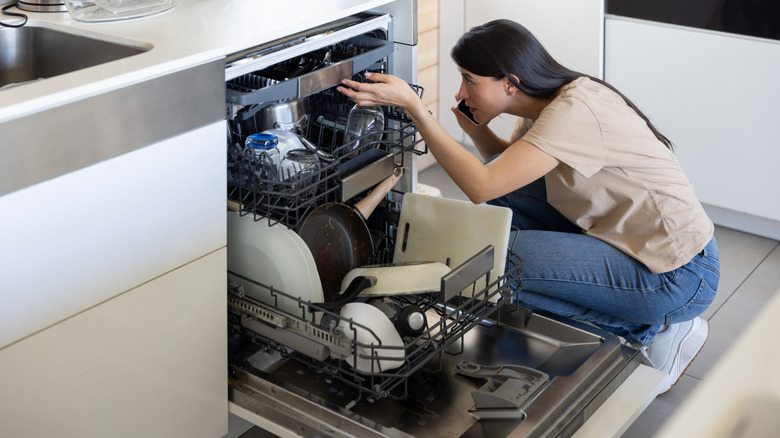 A woman on the phone kneeling beside an open dishwasher.
