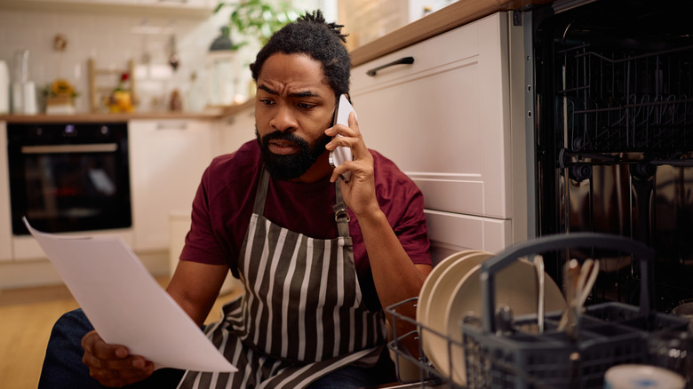 A man on a phone sitting on the kitchen floor next to a dishwasher and looking at a piece of paper.